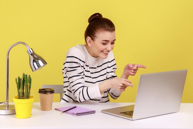 Femme heureuse en chemise rayée pointant vers l'écran d'un ordinateur portable et riant sincèrement en parlant par appel vidéo