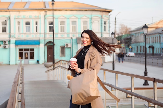 Femme heureuse avec des cheveux ondulés portant un trench-coat bige passant du temps à l'extérieur dans la ville au printemps