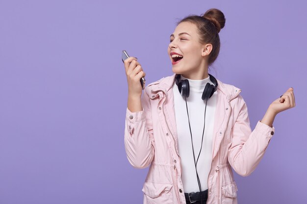 Une femme heureuse avec un chignon apprécie la fin heureuse de la journée, écoute de la musique joyeuse dans les écouteurs, tient le téléphone et chante la chanson, vêtue d'une veste rose pâle et d'une chemise blanche, a des écouteurs autour du cou.