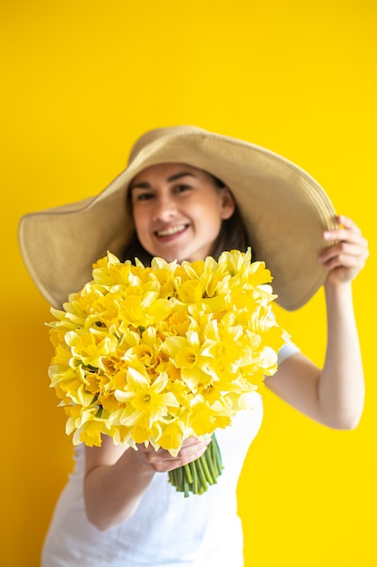 Une femme heureuse dans un chapeau avec un tas de jonquilles jaunes sur mur jaune