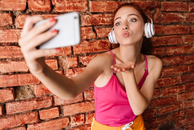femme heureuse dans les écouteurs prenant une photo de selfie sur un téléphone portable tout en envoyant un baiser d'air isolé sur un mur de briques à l'intérieur