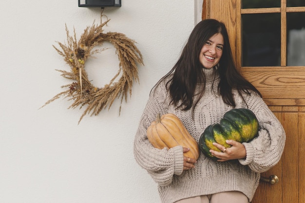 Femme heureuse dans un pull tricoté tenant de grandes citrouilles d'automne pour le décor de l'entrée de la maison Femme élégante décorant la porte d'entrée d'une ferme