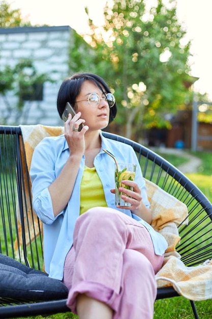 Femme heureuse dans des verres parlant au téléphone portable assis sur une chaise à l'extérieur de l'arrière-cour