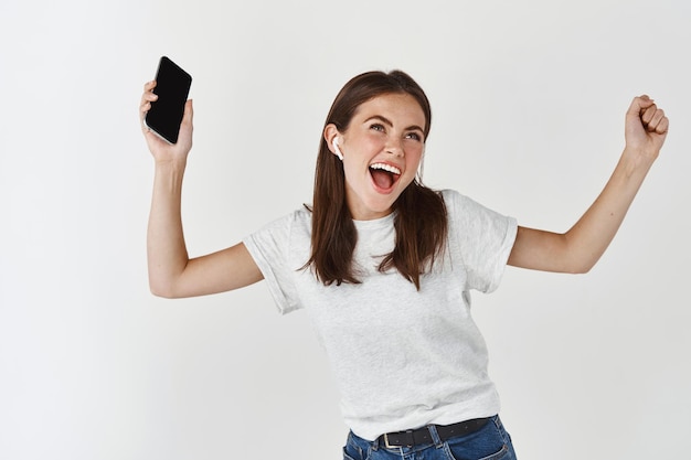 Femme heureuse écoutant de la musique dans des écouteurs sans fil et tenant un smartphone, dansant et chantant sur un mur blanc