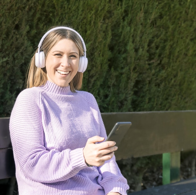 Une femme heureuse écoute de la musique avec des écouteurs sans fil assis sur un banc de parc à l'aide de son smartphone