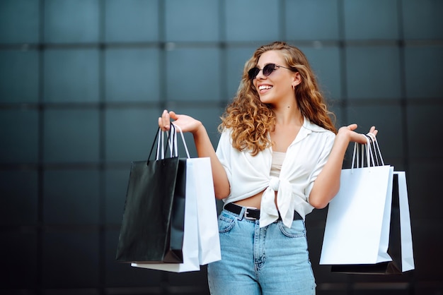 Une femme heureuse et élégante avec des sacs d'achat apprécie le temps ensoleillé en marchant dans la rue pour faire ses courses d'été.