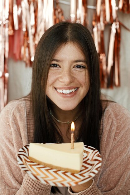 Femme heureuse élégante tenant un morceau de gâteau d'anniversaire avec une bougie à la guirlande de glands roses dans la chambre