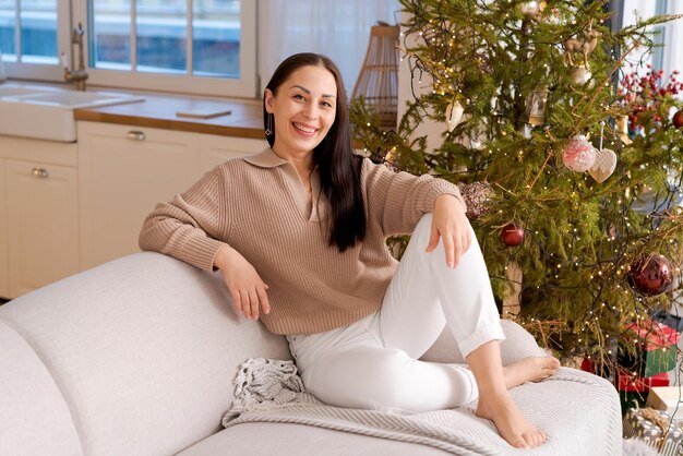 Une femme heureuse est assise sur le canapé en pull à côté d'un arbre de Noël naturel.