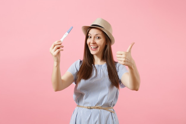 Femme heureuse excitée en robe bleue, tenue de chapeau à la main, regardant le test de grossesse isolé sur fond rose. Soins médicaux gynécologiques, concept de maternité fertilité grossesse. Espace de copie.