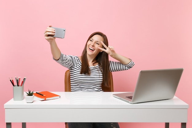 Femme heureuse faisant prise de selfie sur téléphone portable montrant le signe de la victoire tout en s'asseyant et travaillant au bureau blanc avec ordinateur portable pc