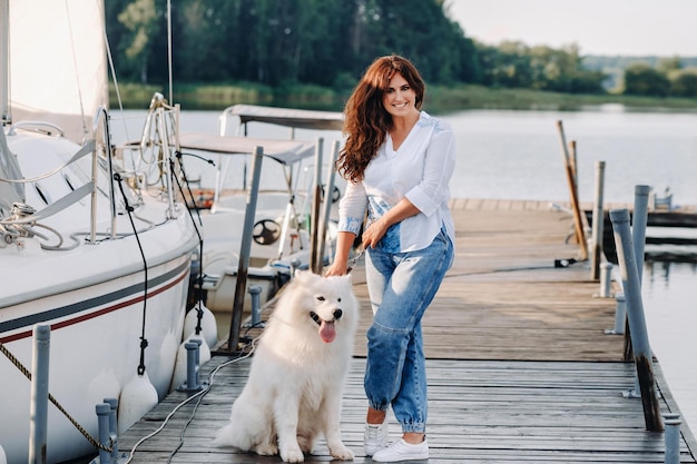 Une femme heureuse avec un grand chien blanc se tient sur la jetée près du yacht