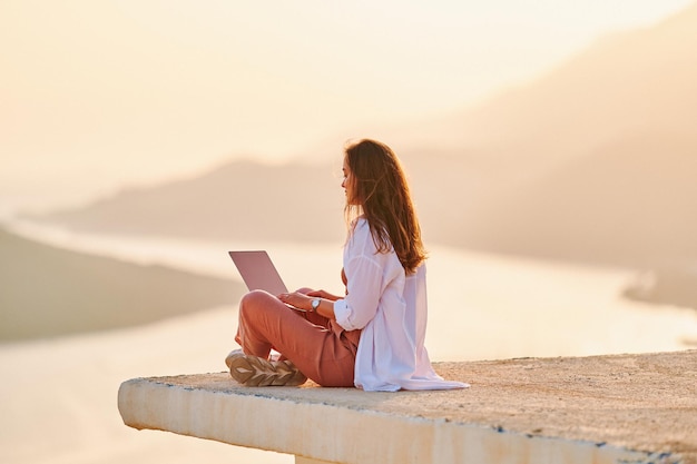 Femme heureuse inspirée assise sur le dessus et profitant de la distance en ligne travaillant à l'ordinateur avec une belle vue au coucher du soleil