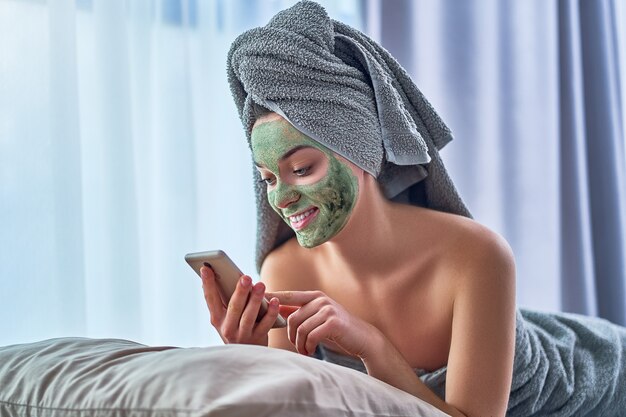 Une femme heureuse et joyeuse avec un masque d'argile nettoyant vert dans une serviette de bain et avec un smartphone se repose et se détend après la douche pendant la journée de spa à la maison. Soins de la peau du visage