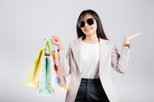 Femme heureuse avec des lunettes shopper sourire tenir des sacs à provisions en ligne multicolores et montrer à la main quelque chose de vide pour le produit actuel, excité femme asiatique achat tourné en studio isolé sur fond blanc