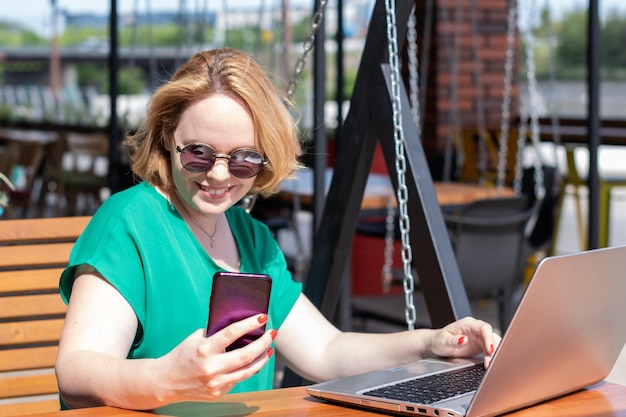 Femme heureuse à lunettes de soleil à l'aide d'une application sur téléphone mobile et ordinateur portable dans un café de rue