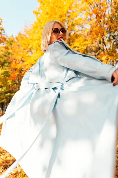 Une femme heureuse avec des lunettes de soleil à la mode dans un élégant manteau bleu se promène dans la nature avec un feuillage d'automne jaune vif