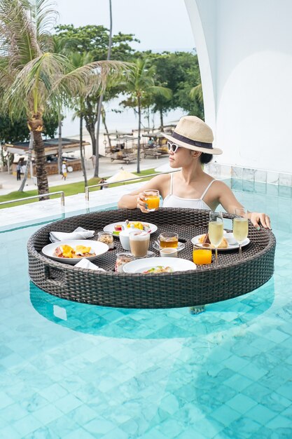 Femme heureuse en maillot de bain blanc ayant un plateau de petit déjeuner flottant dans un hôtel de luxe avec piscine