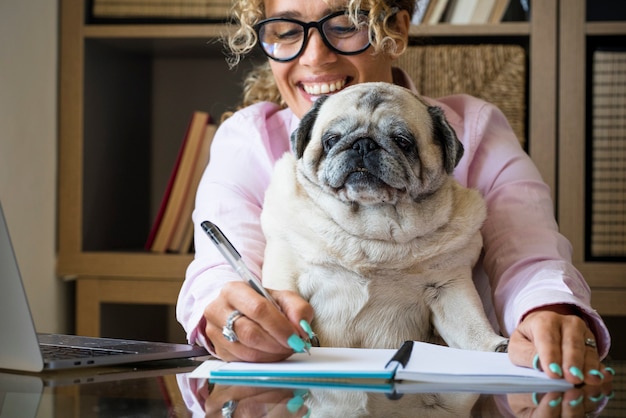 Femme heureuse à la maison amoureuse de son meilleur ami chien carlin tout en travaillant à l'ordinateur portable sur le bureau