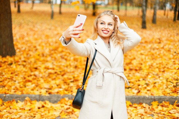 Femme heureuse en manteau tendance avec sac faisant selfie sur smartphone sur fond de feuillage automne jaune