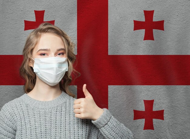 Une femme heureuse avec un masque facial tenant le pouce en l'air sur le fond du drapeau national géorgien Concept d'épidémie de grippe et de protection contre le virus