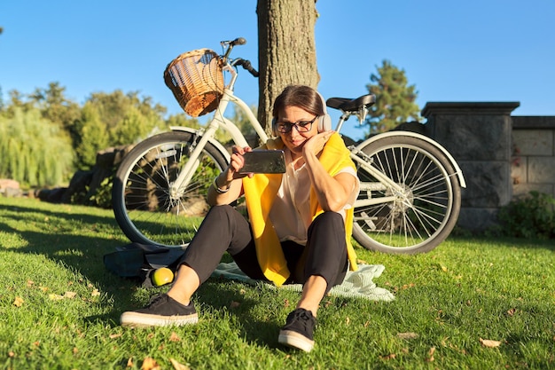 Femme heureuse mature parlant en ligne à l'aide de la communication vidéo. Femme au repos assis dans le parc, avec casque smartphone, fond coucher de soleil bike park