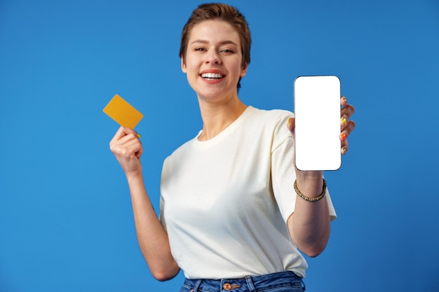 Une femme heureuse montre un écran de smartphone vierge sur fond bleu