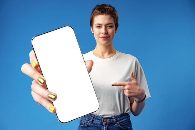 Une femme heureuse montre un écran de smartphone vierge sur fond bleu