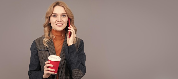 Une femme heureuse parle sur un téléphone portable en buvant du café du matin sur fond gris. Portrait de femme isolée en-tête bannière avec espace de copie