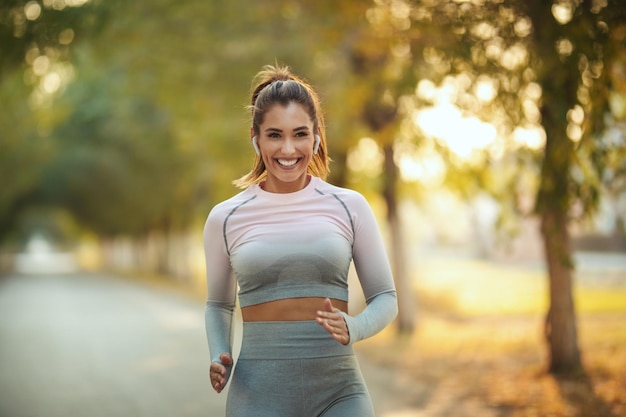 Une femme heureuse, portant des vêtements de sport, avec des écouteurs fait du jogging le long du sentier ensoleillé de l'avenue.