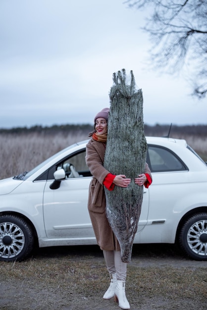 Une femme heureuse porte un arbre de Noël et une voiture nouvellement achetés ou coupés sur le fond en plein air. Concept de préparation au nouvel an. Idée de conte de fées et d'ambiance festive en hiver