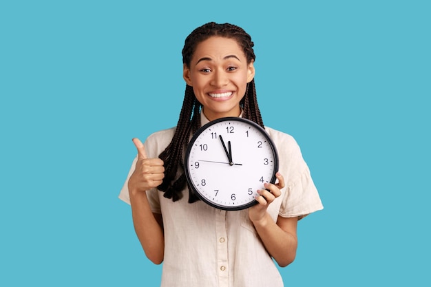 Femme heureuse positive avec des dreadlocks montrant le pouce vers le haut tenant une grande horloge murale regardant la caméra avec un sourire à pleines dents gestion du temps portant une chemise blanche Prise de vue en studio intérieur isolée sur fond bleu