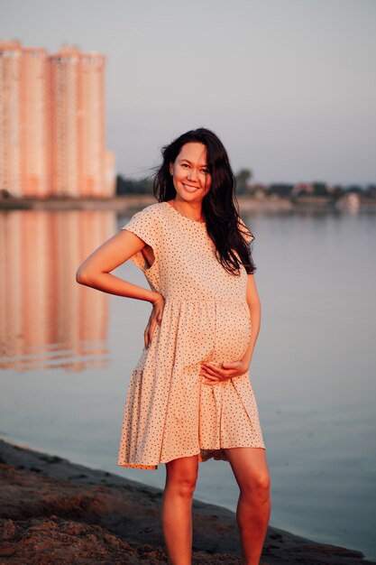 Femme heureuse près des immeubles de grande hauteur enceinte souriante brune asiatique en robe à pois en soie se dresse o...