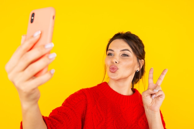 Femme heureuse en pull rouge avec téléphone faisant photo et bisous à la caméra.