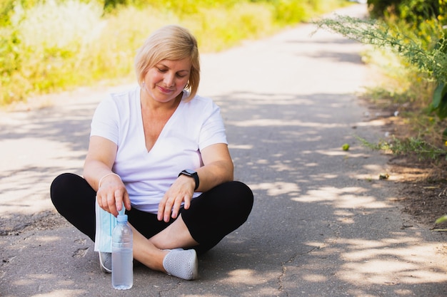 Femme heureuse de remise en forme assise dans la position du lotus sur la route dans le parc seule avec une bouteille d'eau...