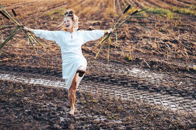 Une femme heureuse en robe danse pieds nus sur un champ boueux tenant des roseaux dans ses mains