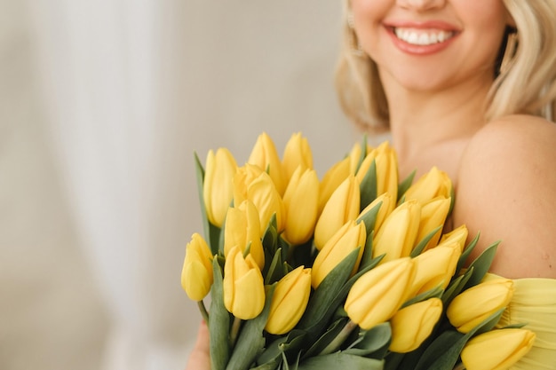 Une femme heureuse en robe jaune embrasse un bouquet de tulipes printanières jaunes à l'intérieur