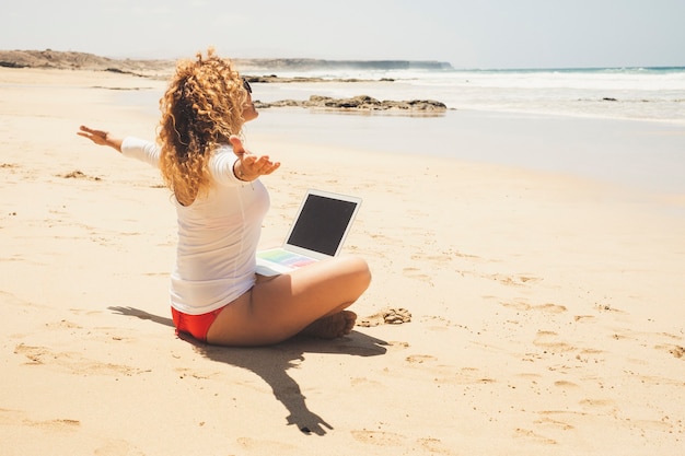 Une femme heureuse s'assied à la plage avec un ordinateur portable sur les jambes