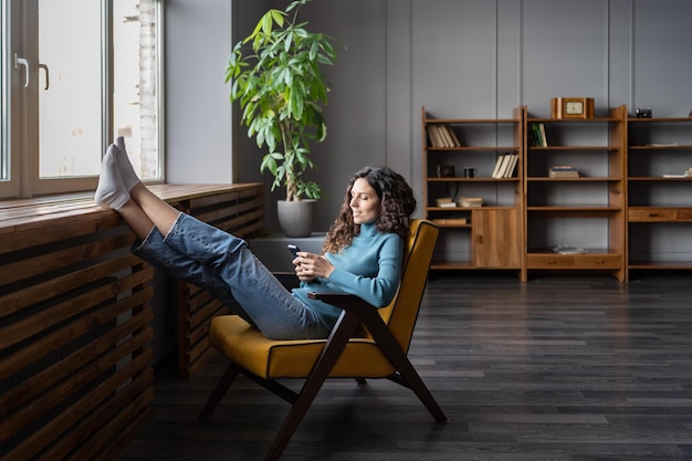 Une femme heureuse se détend dans un fauteuil confortable avec un téléphone portable en utilisant des applications mobiles numériques à la maison