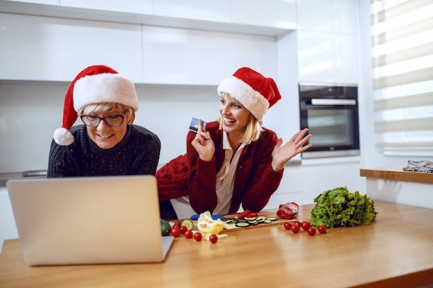 Femme heureuse se penchant sur le comptoir de la cuisine et tenant la carte de crédit. Mère tapant sur ordinateur portable. Les deux ont des chapeaux de père Noël sur la tête. Temps pour les achats de Noël.