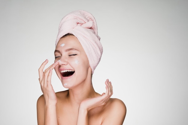 Femme heureuse avec une serviette sur la tête appliquant de la crème sur la peau du visage