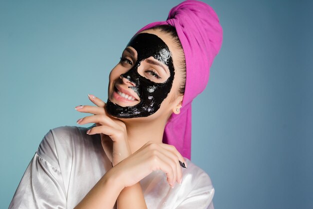 Une femme heureuse avec une serviette sur la tête applique un masque nettoyant sur la peau du visage