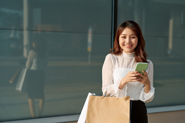 Femme heureuse avec shopping, filles tenant des sacs à provisions à l'aide d'un téléphone intelligent et souriant tout en se tenant à l'extérieur en faisant du shopping