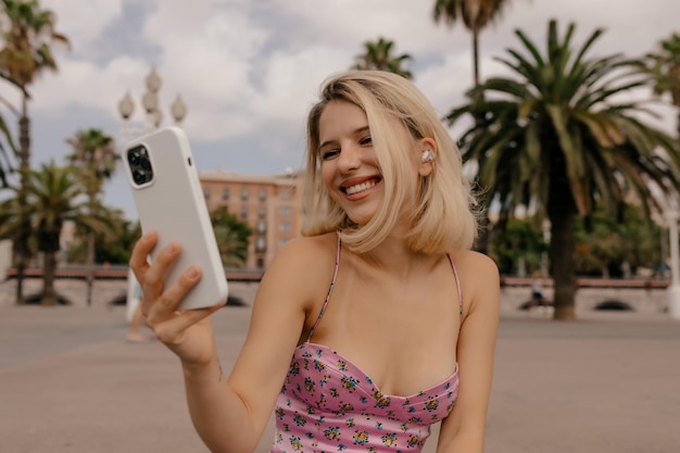 Femme heureuse souriante avec une coiffure courte blonde portant une robe élégante d'été faisant un appel vidéo