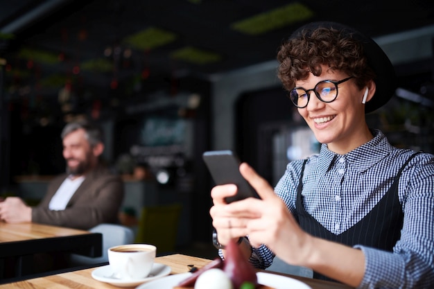 Femme heureuse avec un sourire à pleines dents défilant dans son smartphone tout en passant du temps dans un café confortable