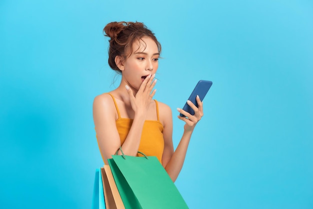 Une femme heureuse surprise tient des sacs en papier bon marché regarde l'écran du téléphone lire des sms vendre un magasin de promotion isolé sur un motif bleu