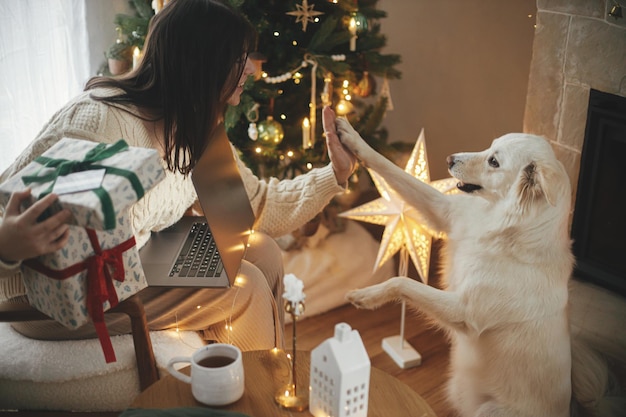 Femme heureuse tenant des cadeaux de Noël élégants avec carte de crédit et jouant avec un joli chien blanc dans une salle de Noël décorée de manière festive avec des lumières, achats de Noël en ligne et ventes du vendredi noir
