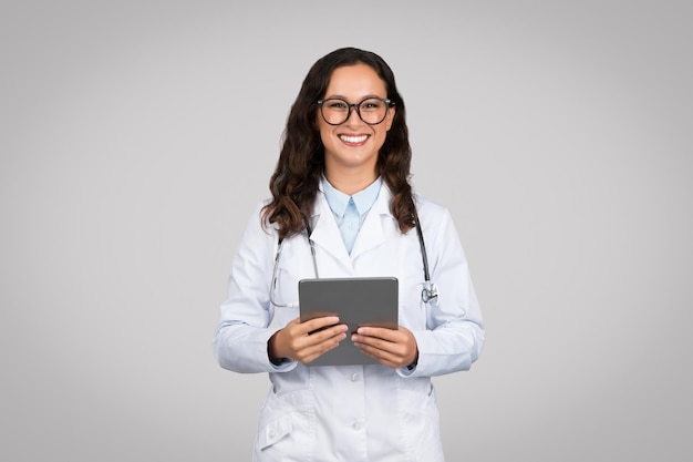 Une femme heureuse tenant une tablette numérique en train de discuter avec des patients et en souriant à la caméra en gris