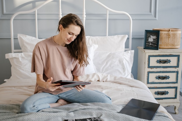 Femme heureuse en tenue décontractée avec les écouteurs travaillant sur un ordinateur portable à distance de la maison sur le lit