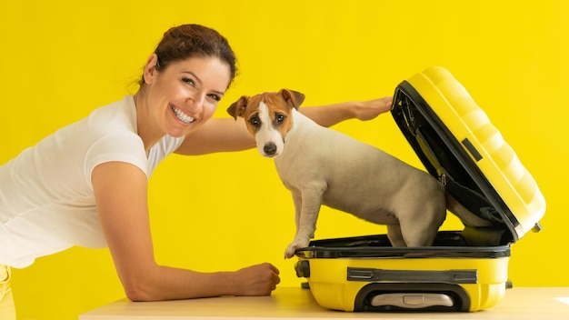 Une femme heureuse tient une valise ouverte avec un chien à l'intérieur sur un fond jaune. Un chiot de Jack Russell Terrier est assis dans un sac à bagages et est prêt à voyager.