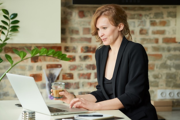 Une femme heureuse travaillant à distance sur un ordinateur portable dans une cuisine. Une dame blonde avec un café discutant d'un projet avec des collègues lors d'une vidéoconférence à la maison.